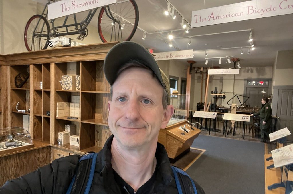 Author taking selfie inside Wright Cycle Company bicycle museum showing historic bicycle displays, workshop exhibits, vintage bicycles hanging overhead, and wooden storage shelving with bicycle components