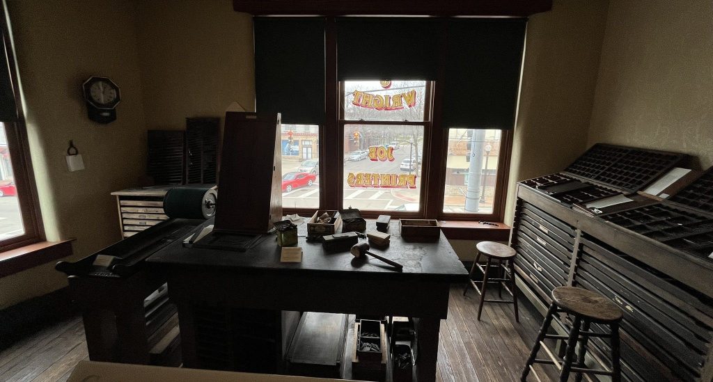 Recreated Wright Brothers printing office showing period printing press, type cases, work desk with printing materials, and window displaying Dayton Tattler newspaper signage