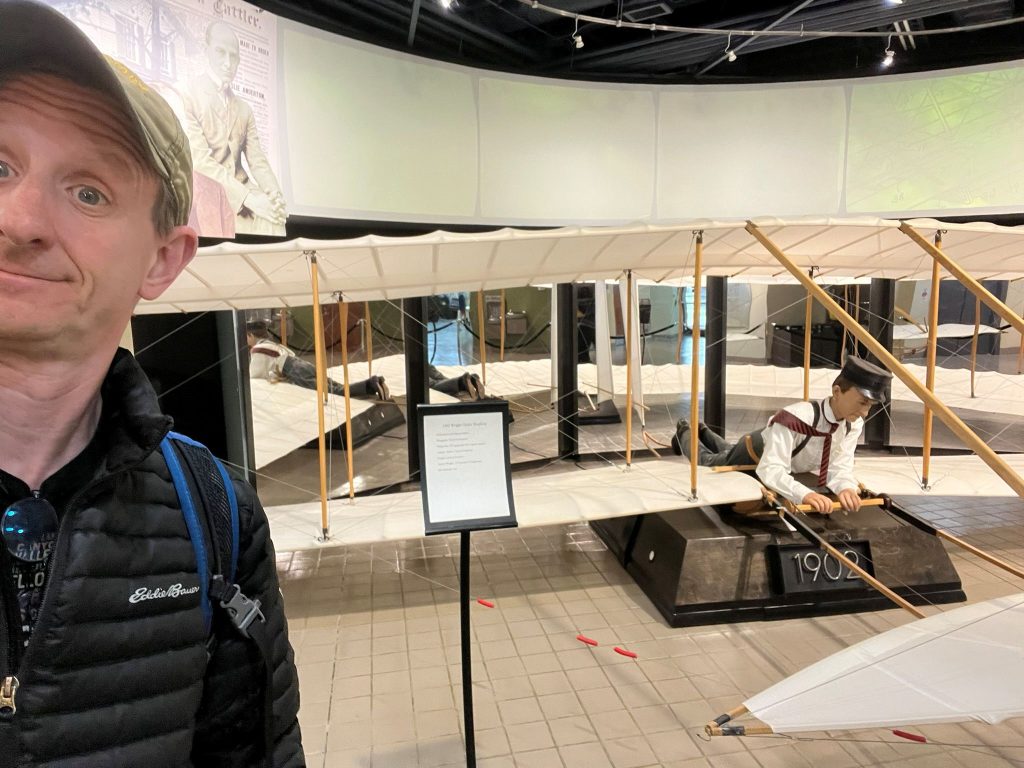 Author taking selfie with Wright Flyer replica display at Dayton Aviation Heritage showing life-size aircraft reproduction with wooden frame, fabric wings, and mannequin figure demonstrating Wright Brothers' historic achievement