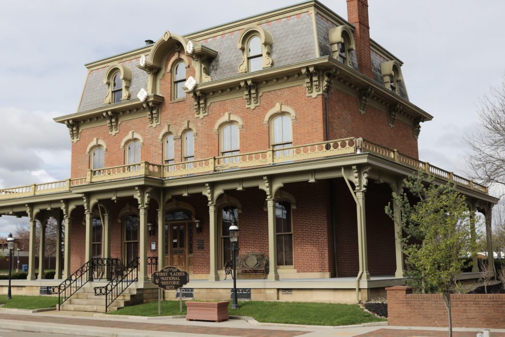 Ohio National Parks: Your Complete Visitor Guide (2026) 7 A wide-angle shot of the First Ladies National Historic Site in Canton, Ohio. The Saxton-McKinley House is a three-story, red brick Victorian-style home featuring a mansard roof with ornate dormer windows and a large, wrap-around wooden porch supported by decorative pillars. A small dark sign in the foreground identifies the location as the "First Ladies National Historic Site," set against a manicured green lawn and a brick sidewalk under a bright, overcast sky.