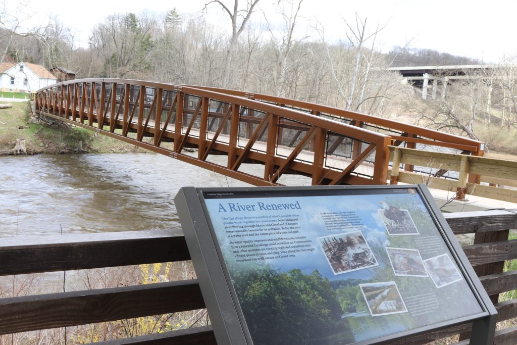 Ohio National Parks: Your Complete Visitor Guide (2026) 8 nterpretive sign titled 'A River Renewed' at Boston Mill Visitor Center overlooks the Cuyahoga River with a distinctive brown wooden boardwalk structure spanning across the water. Bare deciduous trees line both banks, with historic buildings visible in the background on the left hillside
