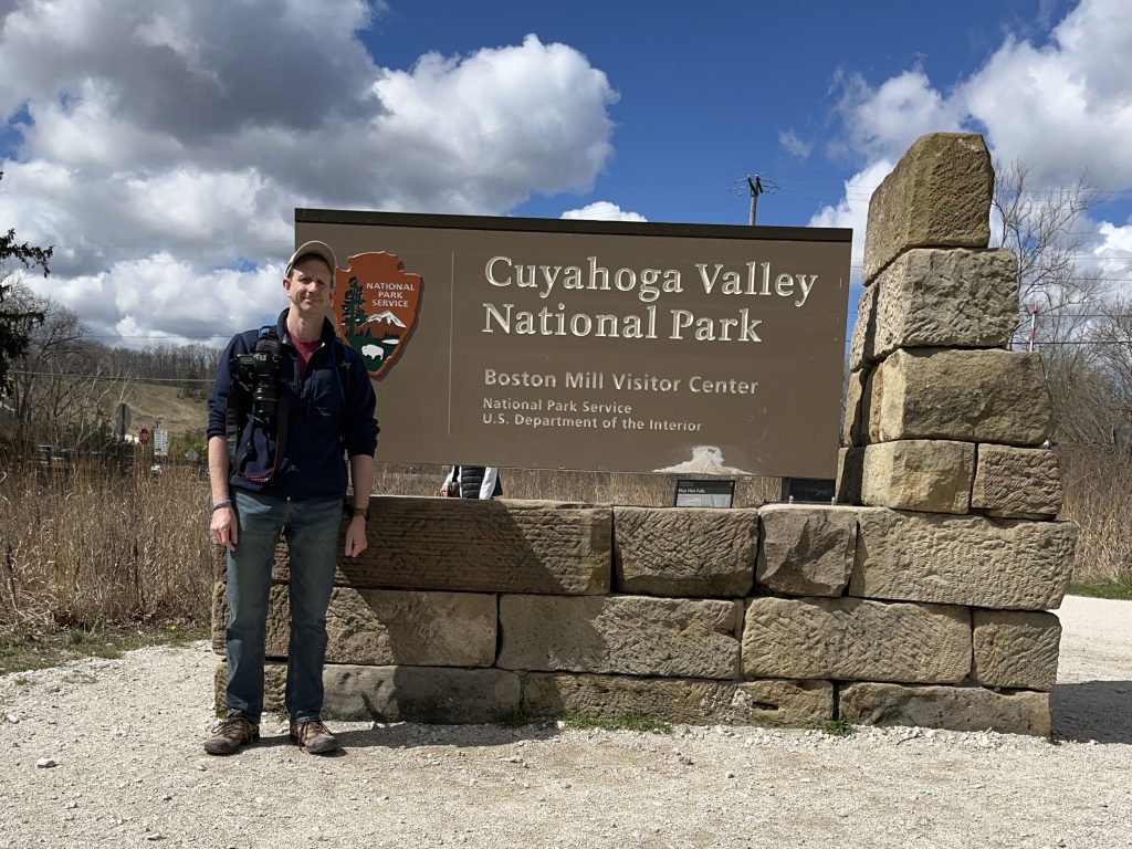 Ohio National Parks: Your Complete Visitor Guide (2026) 1 A man stands smiling next to the large entrance sign for Cuyahoga Valley National Park at the Boston Mill Visitor Center. He is wearing a blue jacket, jeans, and a baseball cap, with a professional camera strapped to his chest. The background features a bright blue sky with fluffy white clouds and the rustic stone architecture of the park sign.