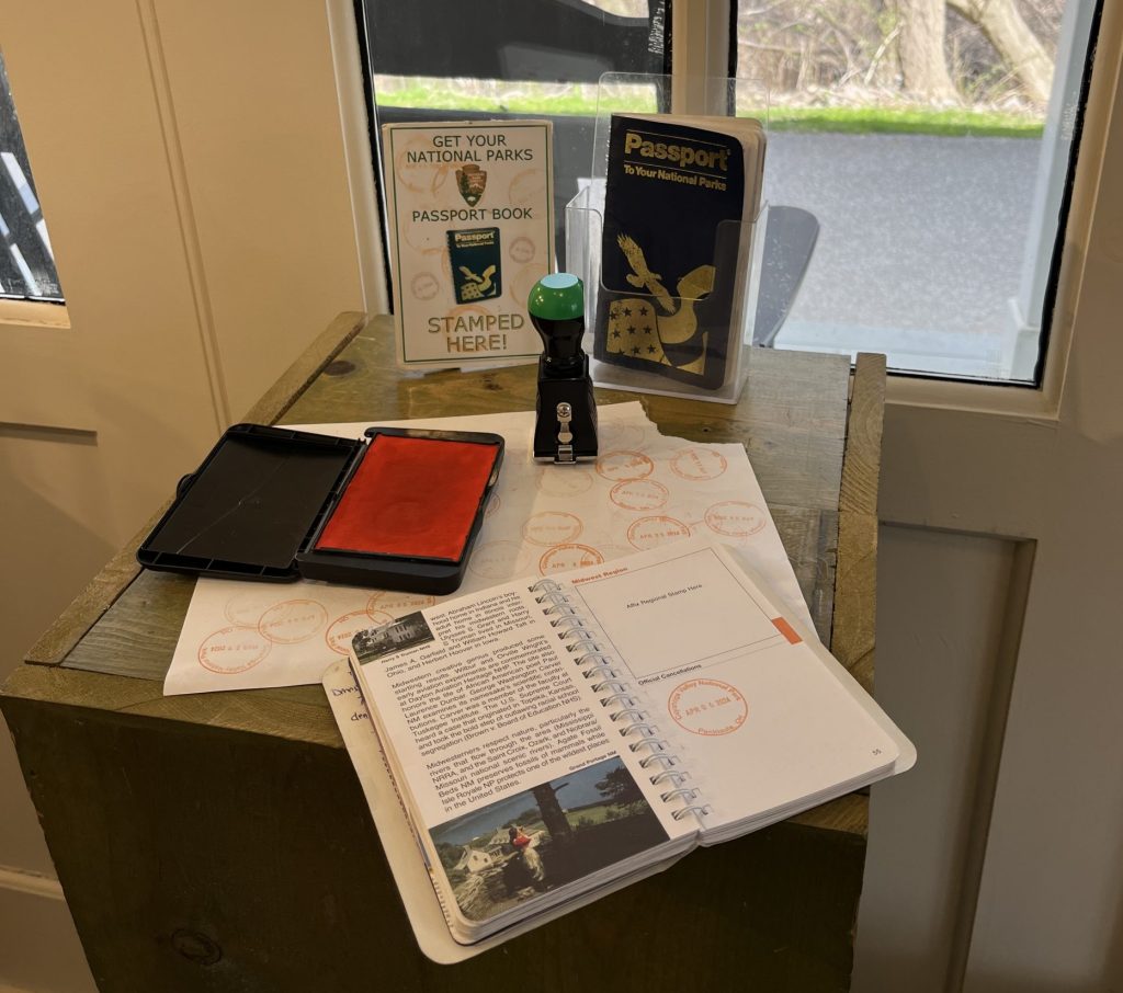 Ohio National Parks: Your Complete Visitor Guide (2026) 9 National Parks passport stamping station at Boston Mill Visitor Center, featuring an open passport book with stamps and photos, two passport covers (one tan with NPS arrowhead logo, one black with golden eagle), red and black ink pads, a stamp, and informational cards about the passport program, displayed on a wooden desk by windows overlooking the grounds.