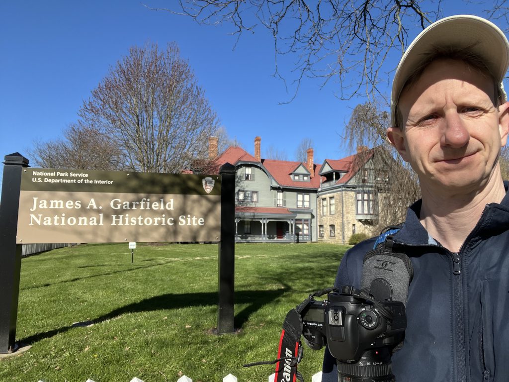Ohio National Parks: Your Complete Visitor Guide (2026) 6 A close-up selfie of a man in a tan baseball cap and blue jacket, smiling while holding a Canon DSLR camera in the foreground. In the background is the James A. Garfield National Historic Site entrance sign, with the official National Park Service and U.S. Department of the Interior logo visible. Behind the sign sits the Victorian-style, multi-gabled "Lawnfield" house with grey siding and a red roof, under a clear blue sky.