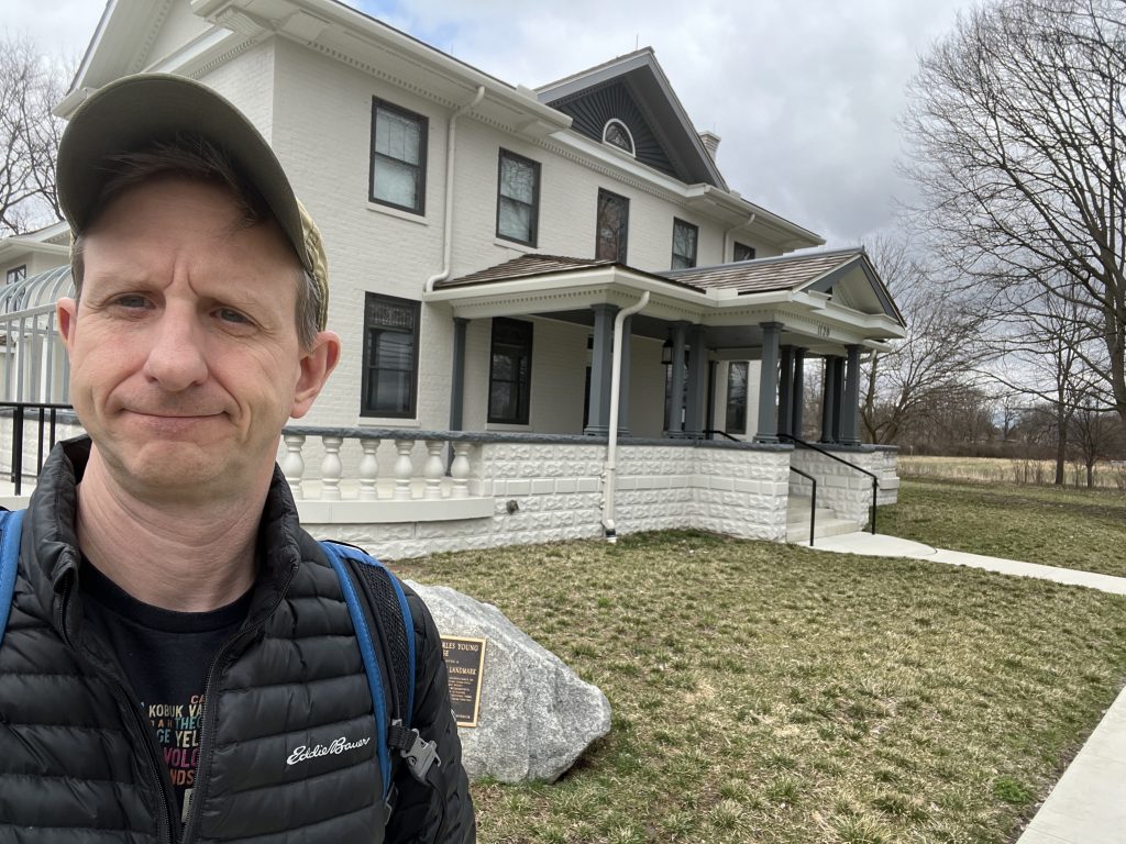 Ohio National Parks: Your Complete Visitor Guide (2026) 4 A man in a black puffer jacket and olive cap stands in front of Youngsholm, the historic white brick home at the Charles Young Buffalo Soldiers National Monument. A large stone with a commemorative bronze plaque is visible in the foreground under a slightly overcast sky.