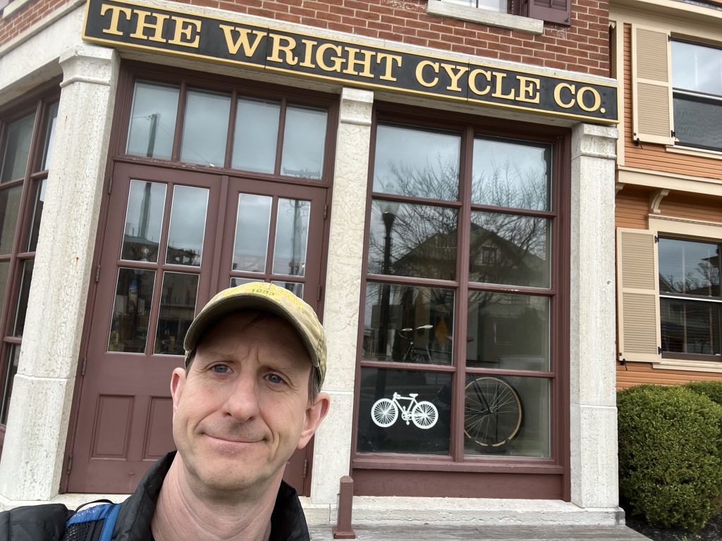 Ohio National Parks: Your Complete Visitor Guide (2026) 3 A selfie of a man in a tan baseball cap standing in front of the historic "The Wright Cycle Co." building, located next to the Dayton Aviation Heritage National Historical Park. The storefront features a red brick exterior, dark red wooden window frames with a white bicycle decal, and a prominent black and gold sign above the entrance.