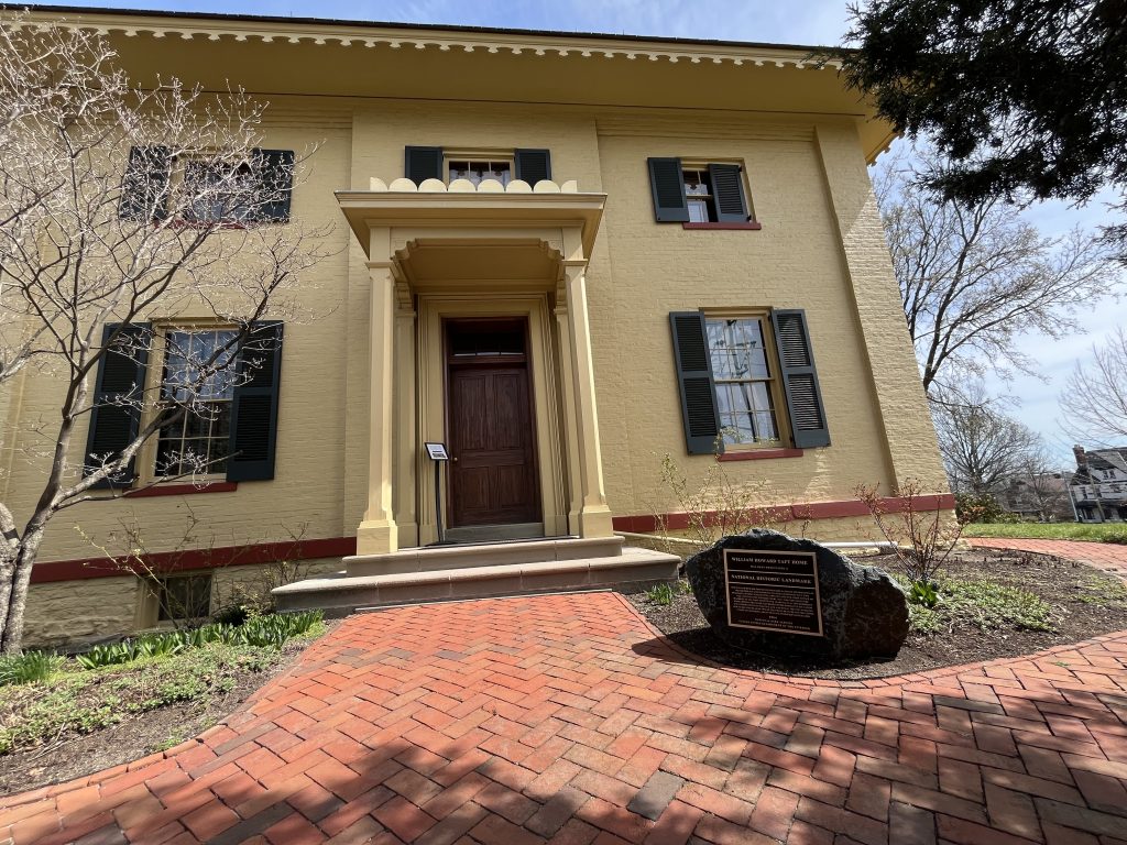 Ohio National Parks: Your Complete Visitor Guide (2026) 5 A wide-angle shot of the William Howard Taft National Historic Site in Cincinnati, Ohio. The two-story, yellow-painted brick home features dark green shutters and a formal wooden front door under a small portico. A red brick herringbone-patterned path leads to the entrance, and a large commemorative rock with a bronze plaque marking it as a National Historic Landmark sits in the landscaped garden to the right.