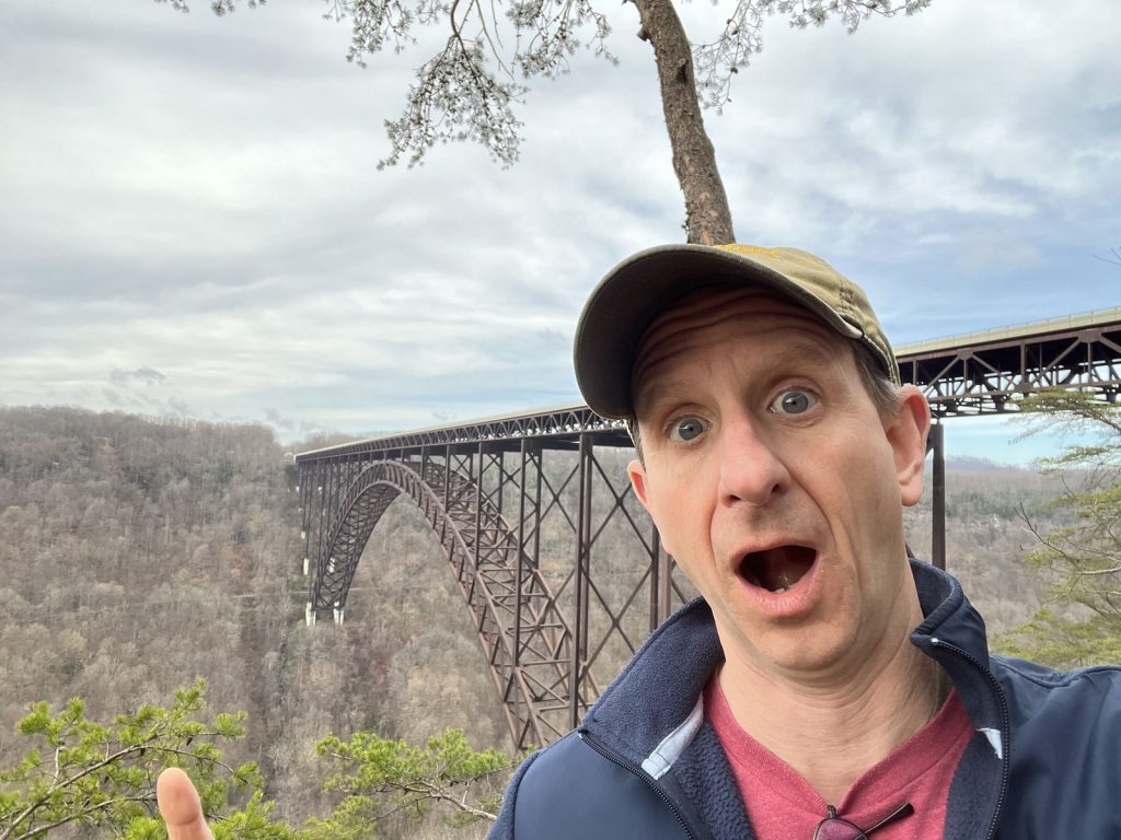 Visitor selfie with exaggerated surprised expression in front of New River Gorge Bridge arch spanning the gorge