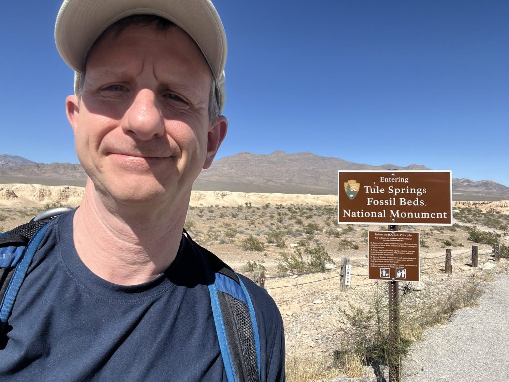 olo visitor selfie at the Entering Tule Springs Fossil Beds National Monument sign with desert badlands and mountains in the background